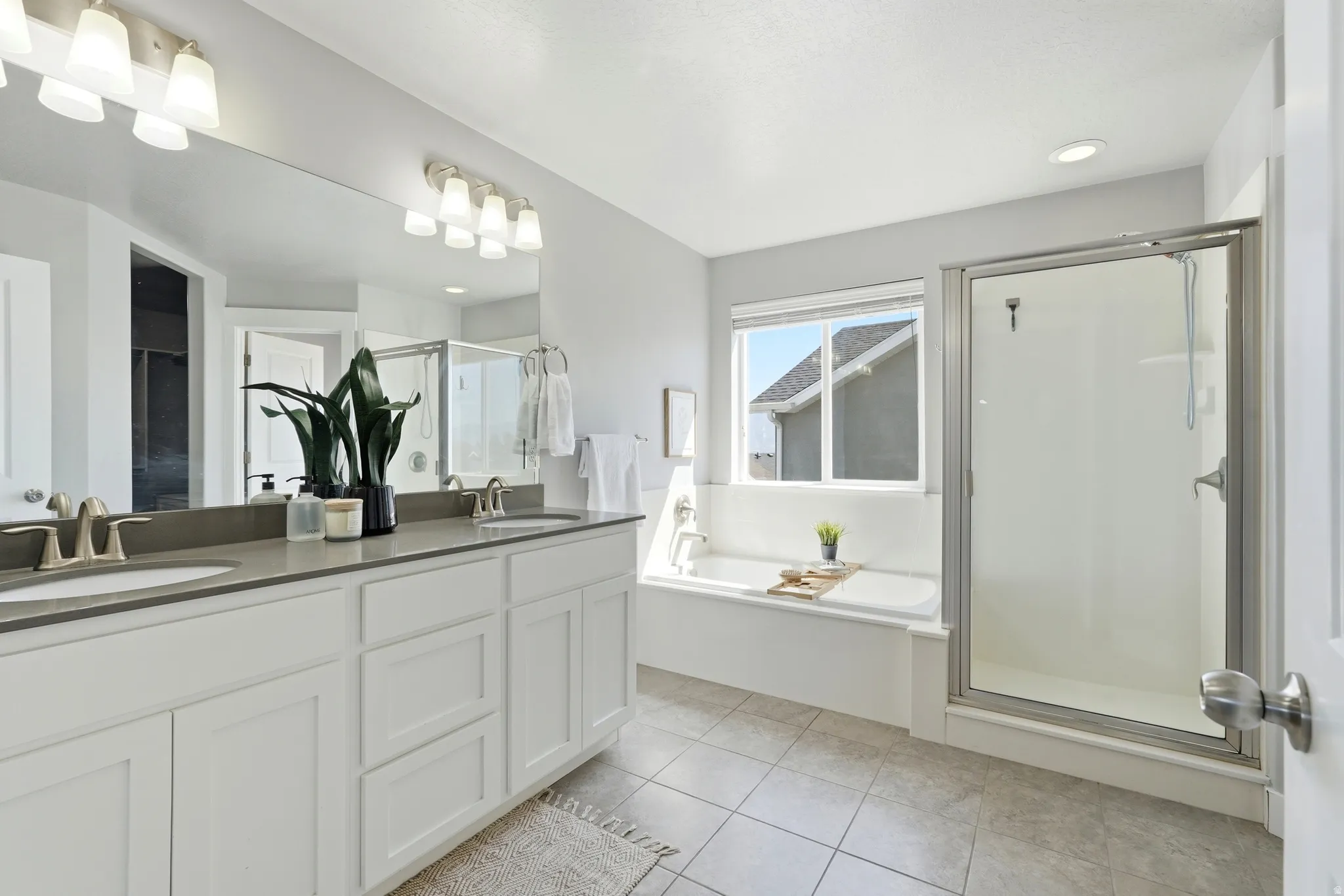 Dual vanity with undermount sinks, full-width mirror, tile flooring, glass-enclosed shower, and built-in tub with window