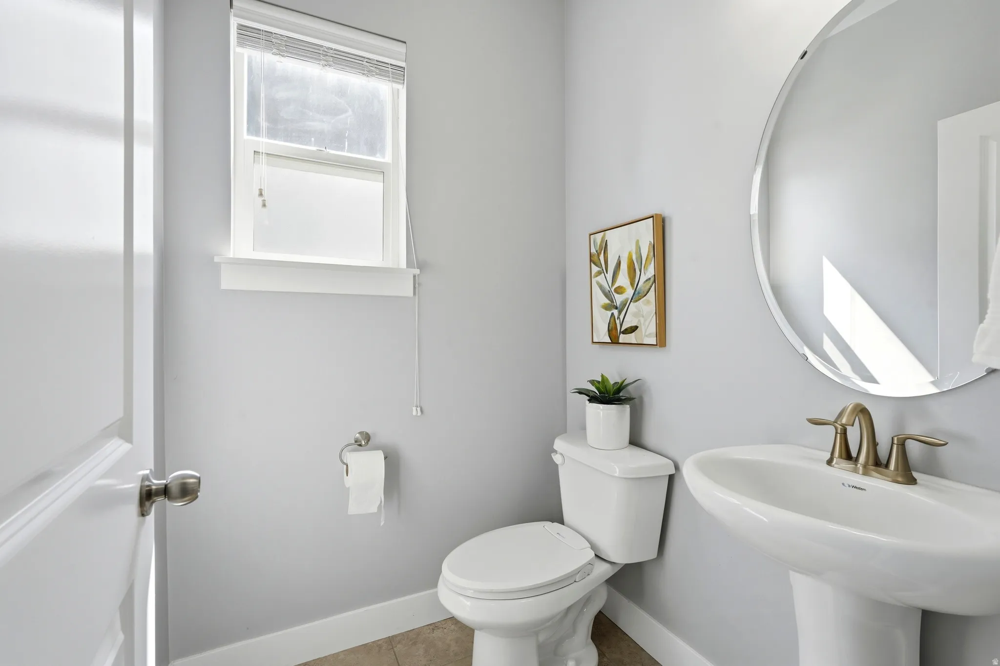 Pedestal sink with a brass-finish faucet, circular wall mirror, and a single window