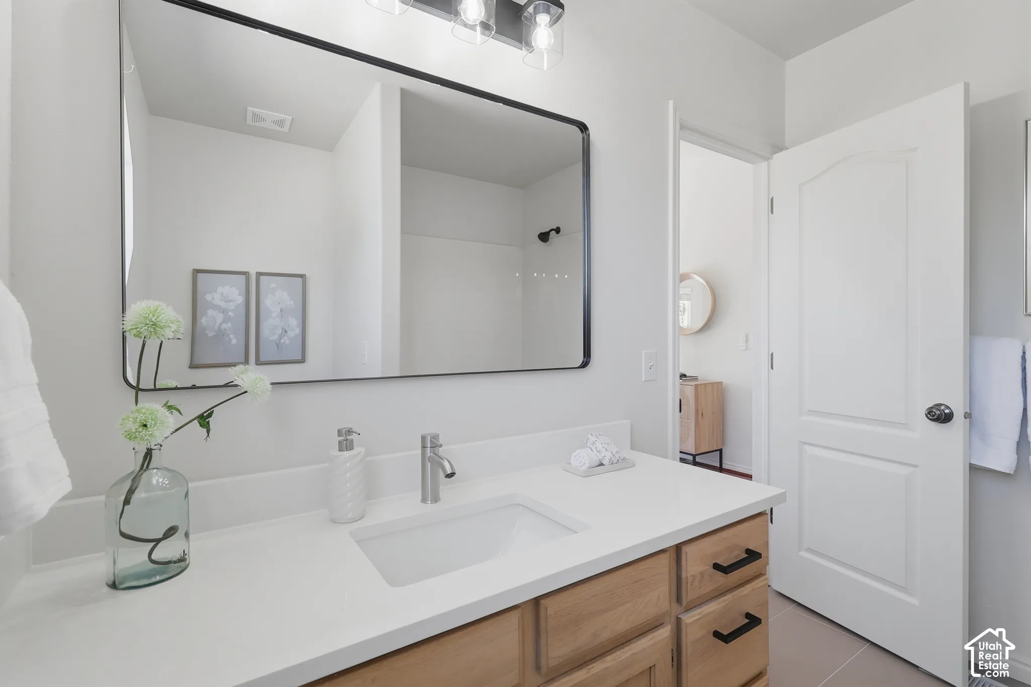 Light wood-finish vanity with a white countertop and integrated sink, complemented by a black-framed rectangular mirror and a three-light vanity fixture