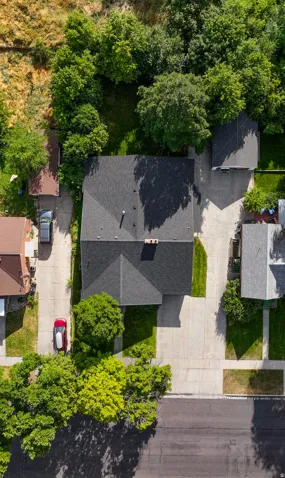 Property featuring a dark shingle roof, paved driveways, a landscaped front yard, and mature trees surrounding the rear