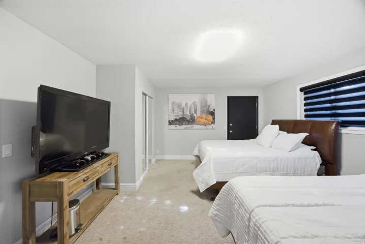 Carpeted flooring, light grey wall paint, dark window blinds, mirrored closet doors, and a dark brown headboard