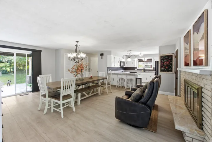 Light wood-finish flooring throughout, brick fireplace with mantel, white kitchen cabinetry with dark countertops, sliding glass door, and a chandelier lighting fixture