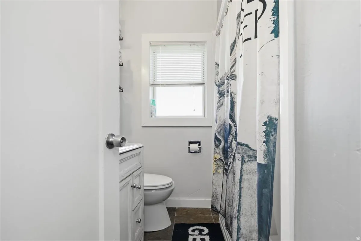 Window with blinds, white vanity with cabinet storage, white toilet, and dark-toned floor tiles