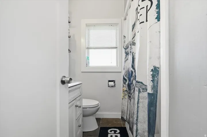 Window with blinds, white vanity with cabinet storage, white toilet, and dark-toned floor tiles