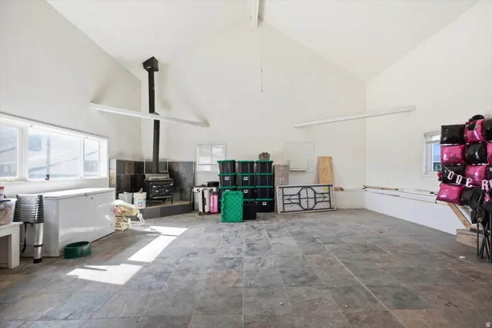 Vaulted ceiling with exposed beams, wood-burning stove with a tiled surround, slate tile flooring, multiple windows, and a built-in workbench