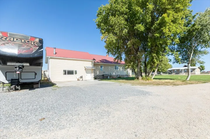 Gravel driveway with ample parking, mature trees, and a building featuring a red metal roof and beige siding