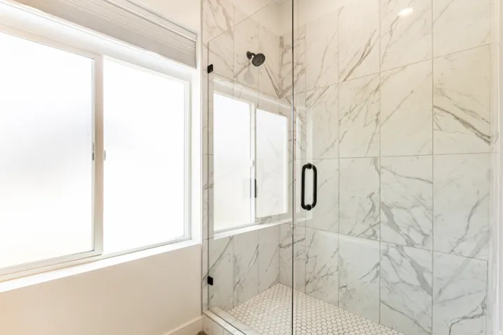 Walk-in shower with clear glass enclosure, matte black fixtures, white marble-look wall tiling, and hexagonal floor tiling