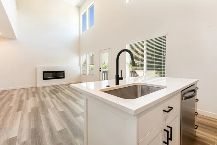 Open-concept living space with vaulted ceilings, large windows, wood-finish flooring, a linear fireplace, and a kitchen island featuring a stainless steel sink, matte black faucet, and white countertop