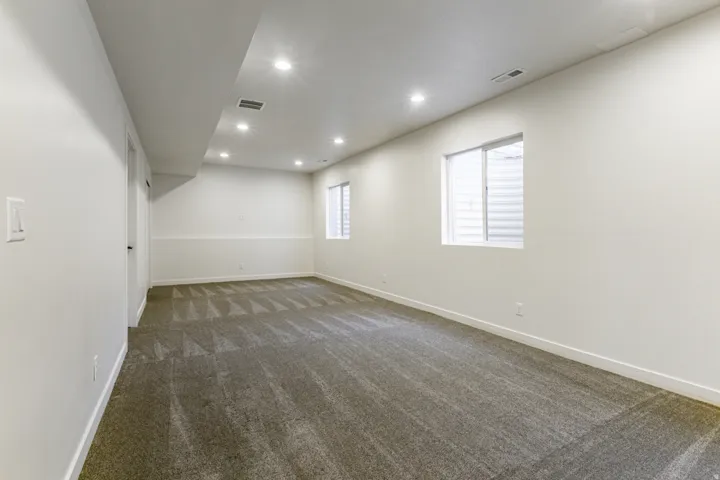 Recessed lighting illuminates the open floorplan, featuring two windows and gray carpeting