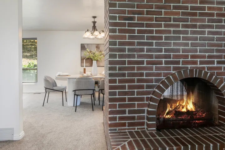 Brick-faced fireplace with an arched opening, light-toned carpeting, and a dining area featuring a modern chandelier