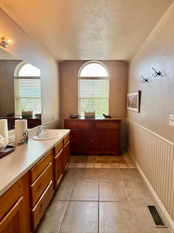 Dual arched windows, single vanity with light wood cabinetry, tile flooring, and wainscoting