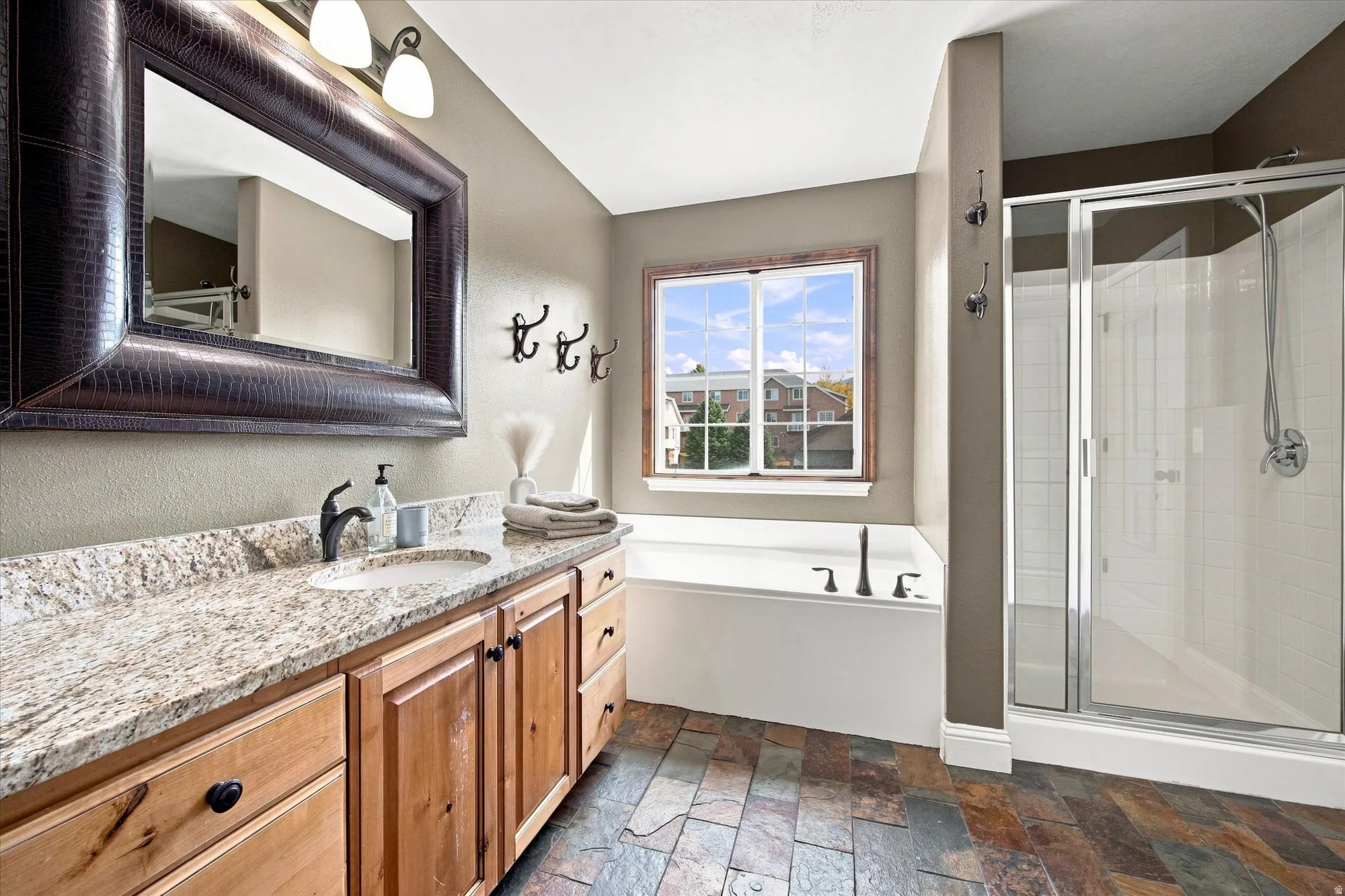 Dual-sink vanity with granite countertop and wood cabinetry, a built-in soaking tub with a window, a walk-in shower with a glass enclosure, and multi-tone slate tile flooring