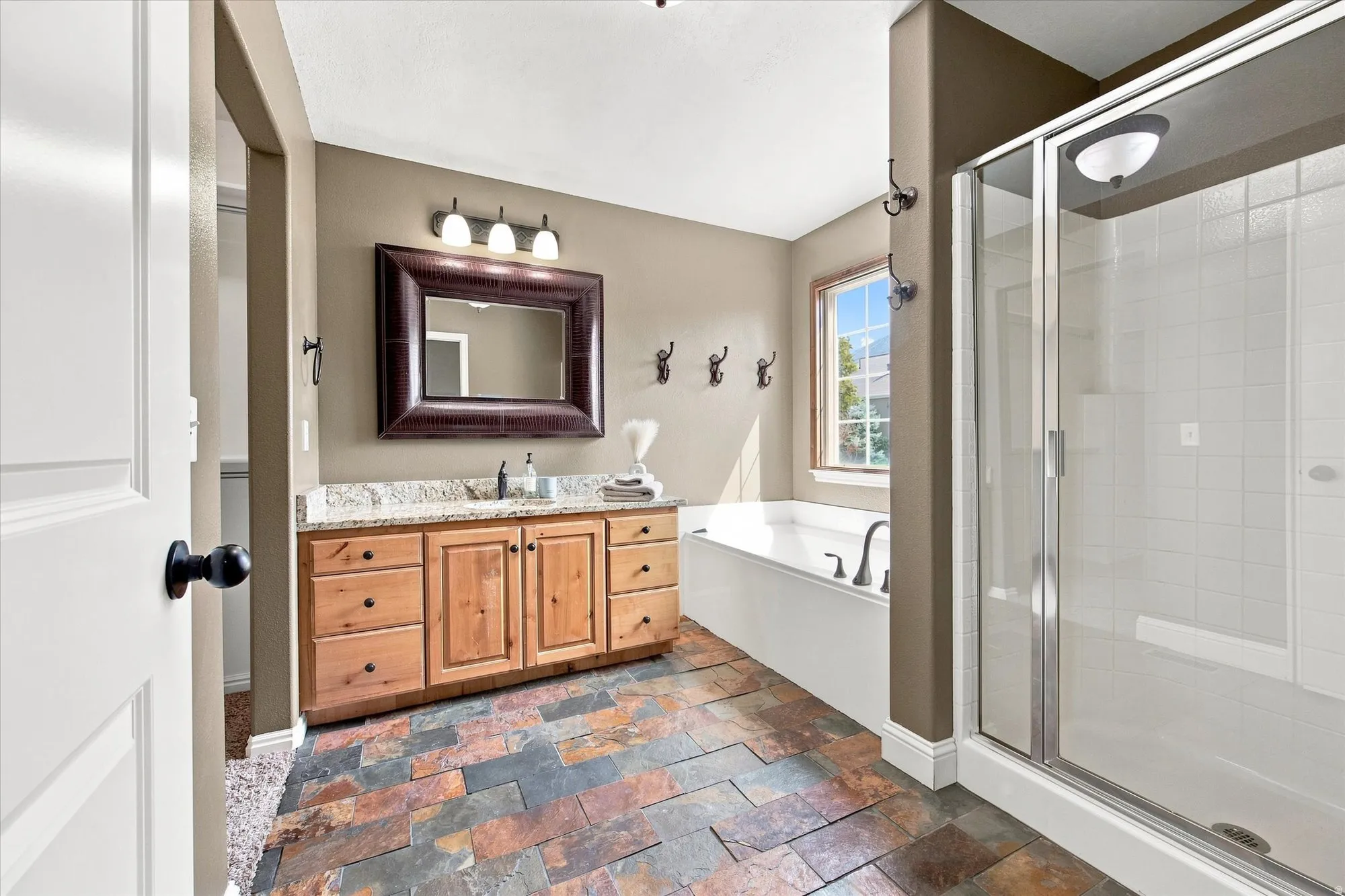 Warm-toned tile flooring, double vanity with light wood cabinetry, stone countertop, built-in bathtub, and a glass-enclosed shower