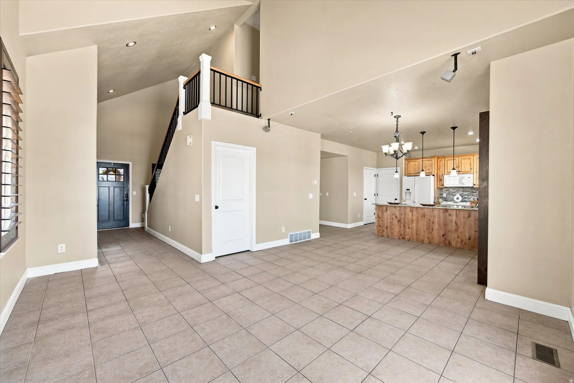 Spacious living area featuring tile flooring, vaulted ceilings, a prominent staircase with black railings, and a kitchen with wood-finish cabinetry and a tiled backsplash