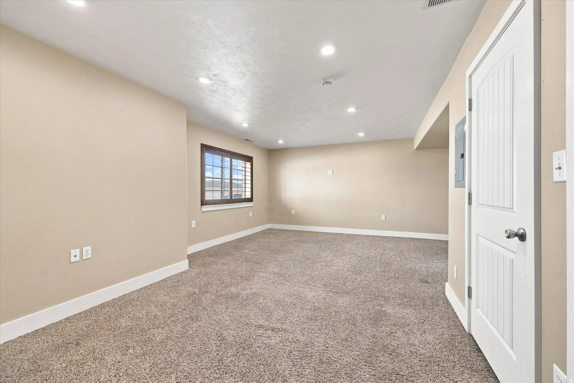 Recessed ceiling lighting, neutral wall paint, window with wood-finish trim, textured carpet flooring, and white paneled door