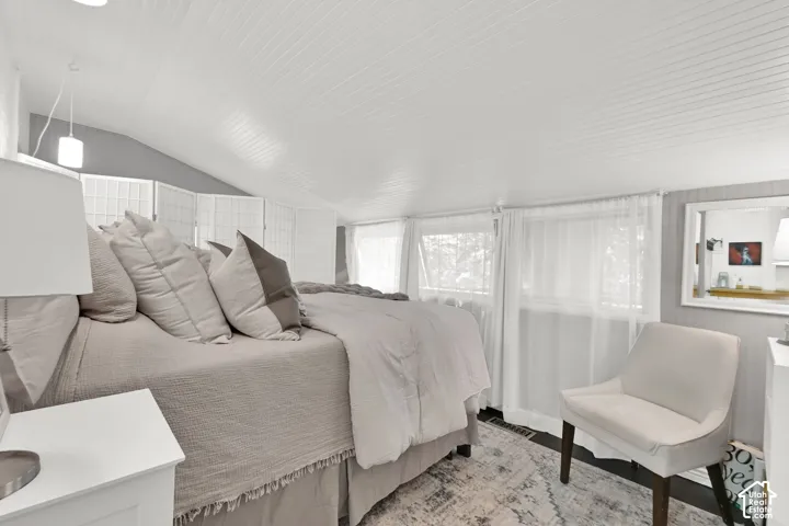 Bright, airy primary bedroom suite featuring angled vaulted ceilings, expansive windows with sheer drapery, hardwood flooring, and an en-suite bathroom with a framed mirror and vanity
