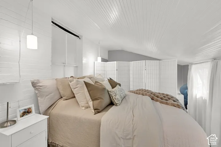 White brick accent wall, sloped wood-paneled ceiling, pendant lighting, white window treatments, and a white nightstand