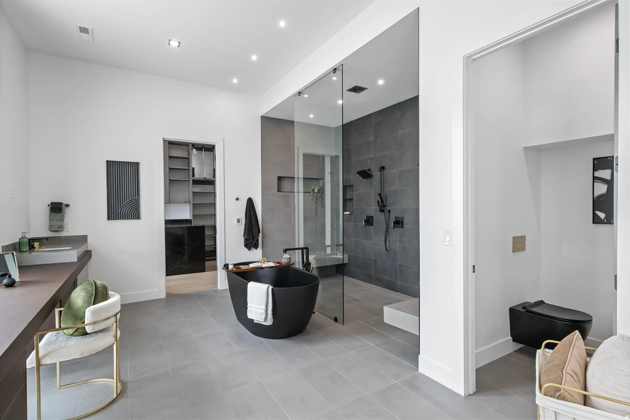 Freestanding matte black soaking tub, enclosed shower with dark tile surround and built-in bench, dark wood-finish vanity with integrated sink, walk-in closet with built-in shelving, and a private toilet area