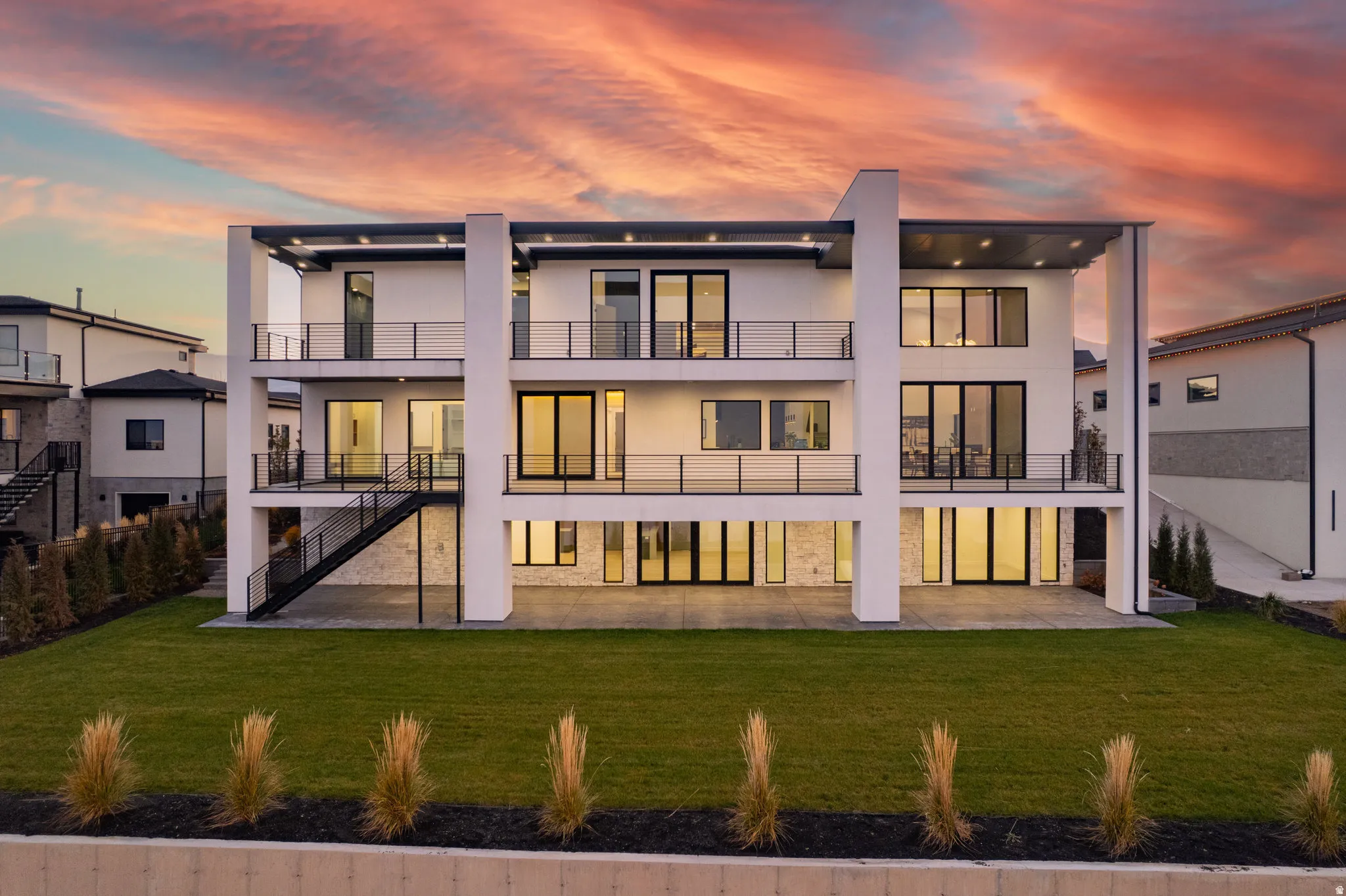 Multi-level exterior with multiple balconies, expansive windows, black railing accents, a lower-level patio, and a manicured lawn