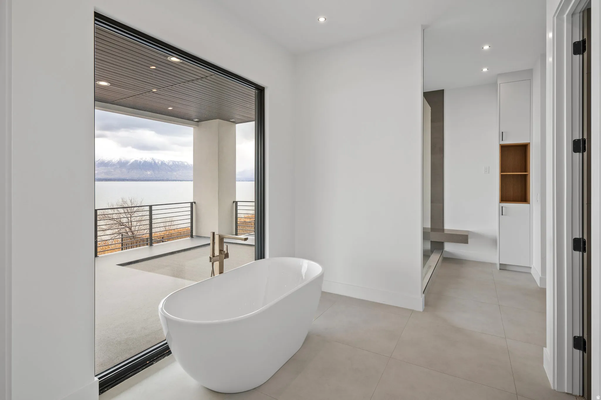 Freestanding soaking tub with floor-mounted faucet, expansive sliding glass doors, private balcony with metal railing, large format tile flooring, and recessed lighting