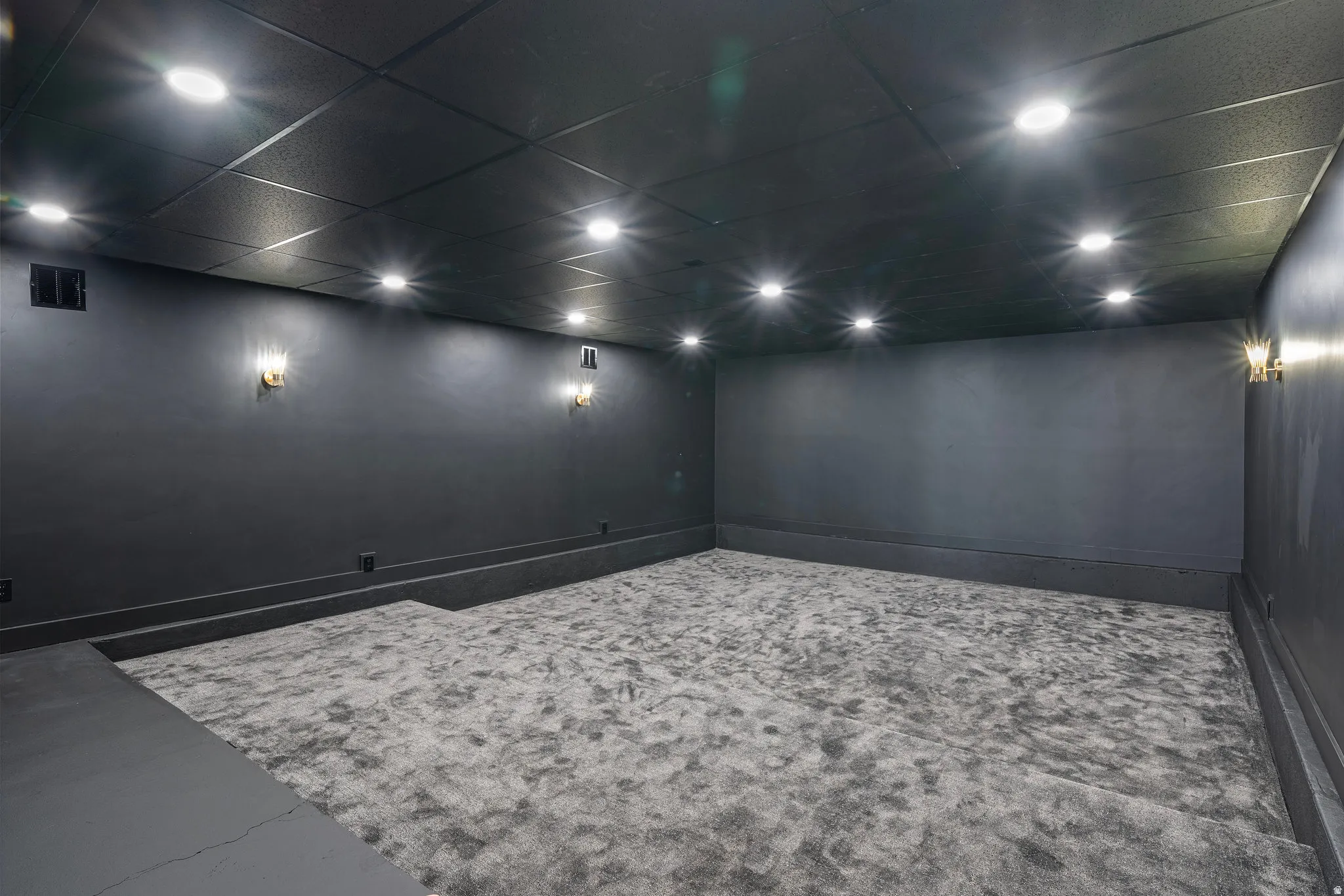Recessed ceiling lighting, wall-mounted sconces, dark painted walls, dark ceiling panels, and light-toned carpet flooring