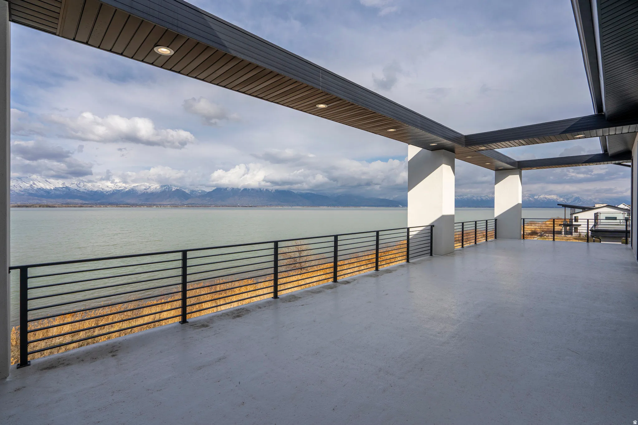 Expansive covered deck featuring a smooth concrete surface and black horizontal railings, overlooking a large body of water with distant snow-capped mountains