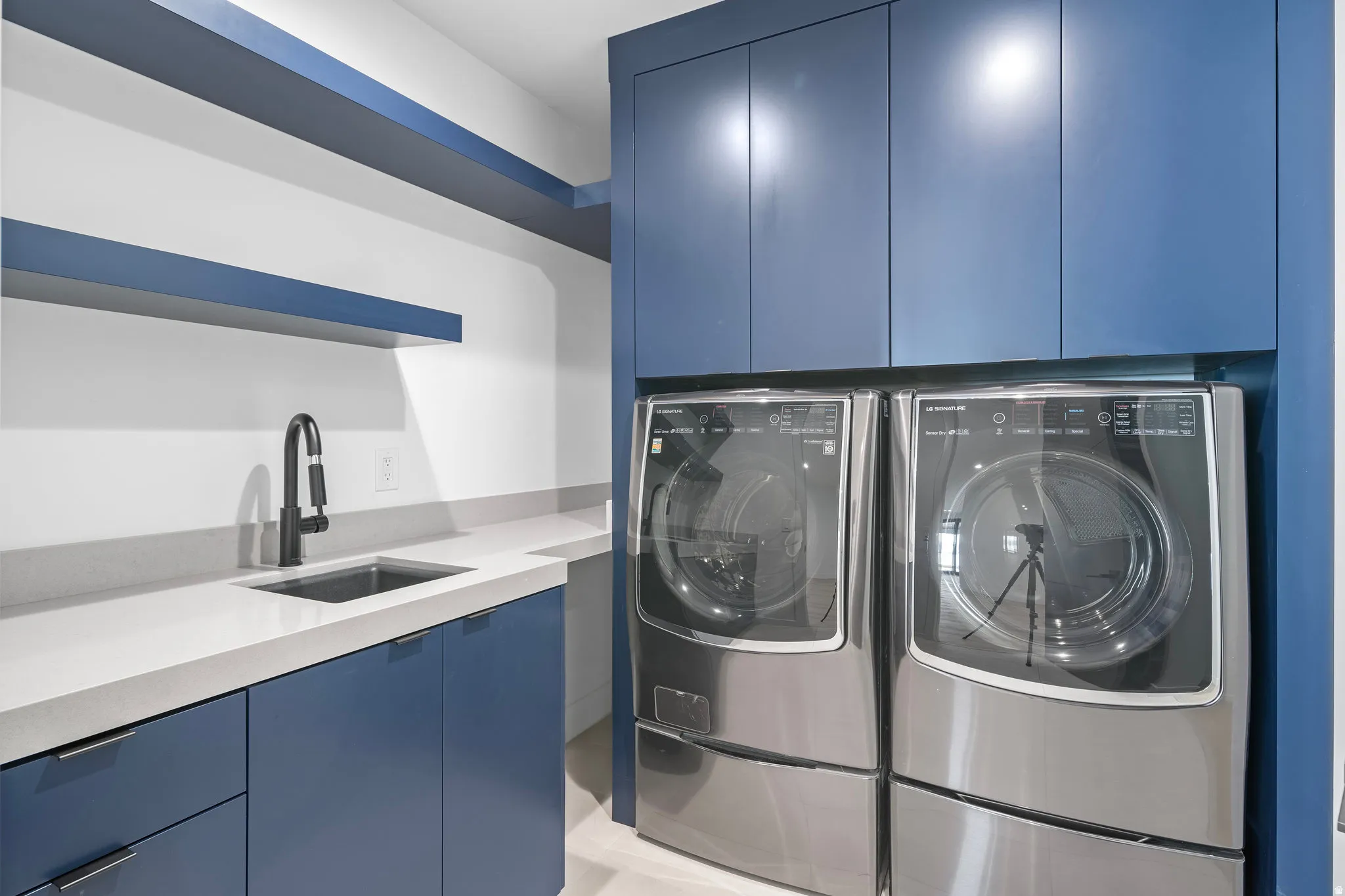 Laundry area featuring rich blue cabinetry, light-toned countertops, a stainless steel sink with a matte black pull-down faucet, and open shelving