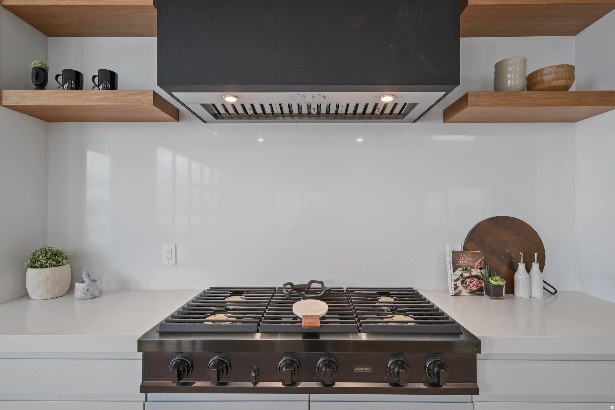 Stainless steel range with black knobs, dark finish range hood, white countertops, white backsplash, and wood-finish floating shelves