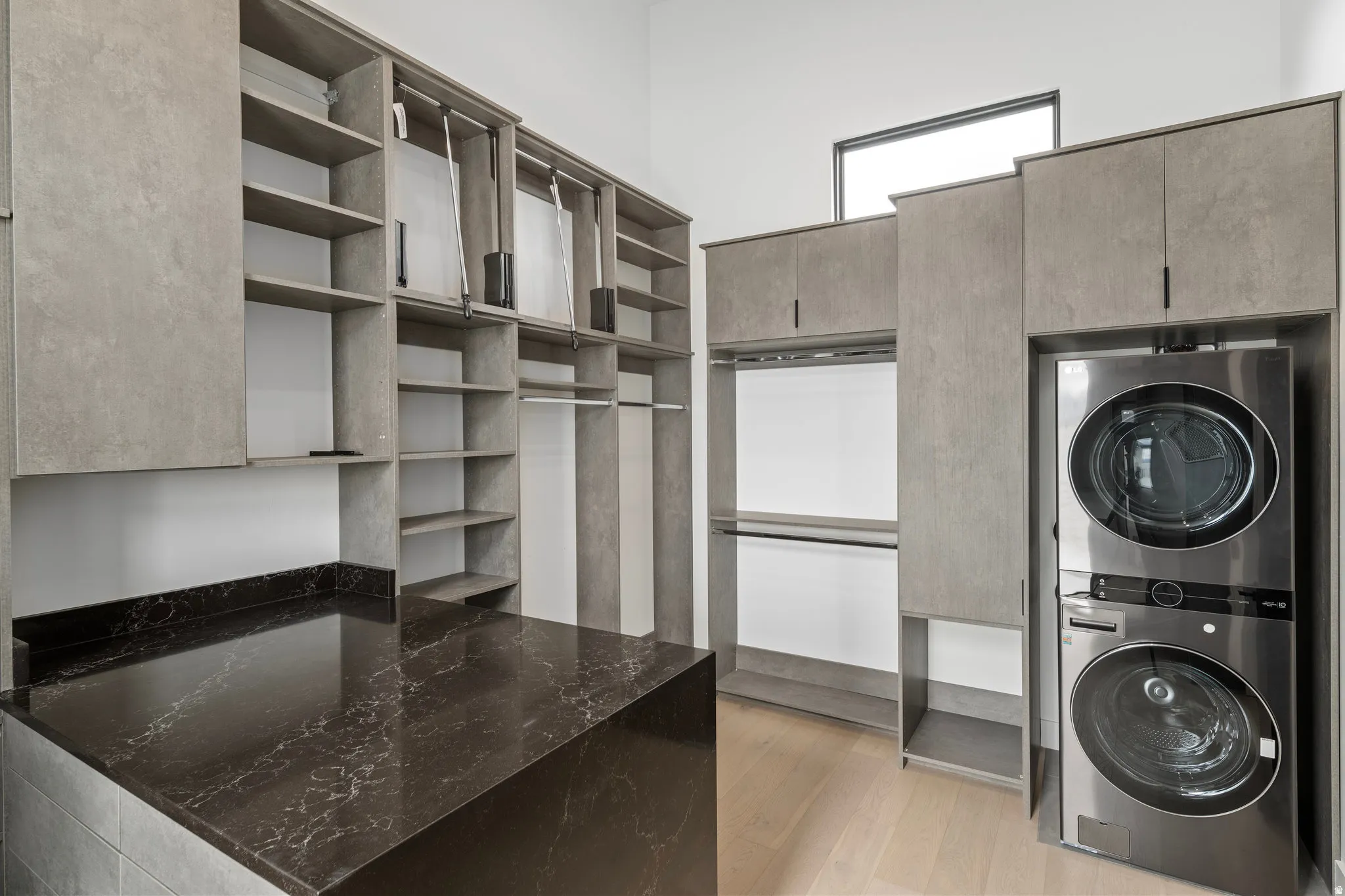 Built-in cabinetry with extensive shelving, integrated clothing rods, a dark stone countertop, stacked stainless steel laundry appliances, and a high window