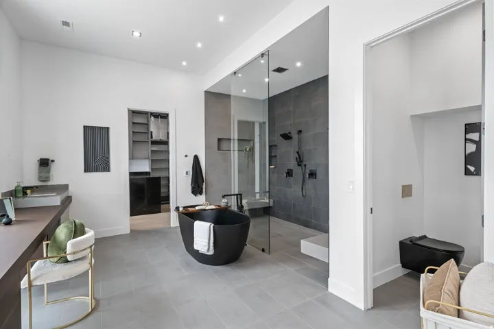 Freestanding matte black soaking tub, enclosed shower with dark tile surround and built-in bench, dark wood-finish vanity with integrated sink, walk-in closet with built-in shelving, and a private toilet area
