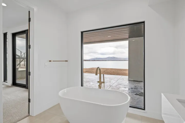 Freestanding soaking tub with a floor-mounted gold-finish faucet, large picture window with mountain scenery, and a white vanity with an integrated basin