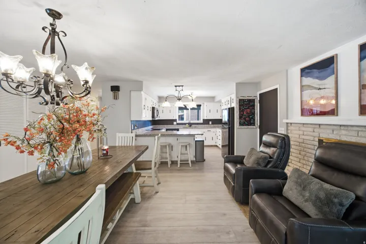 Open-concept living space featuring wood-finish flooring, a stone fireplace, and a kitchen with white cabinetry and dark countertops