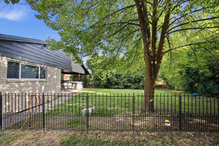 Expansive fenced backyard featuring a manicured lawn, mature trees, and a covered patio area with a hot tub