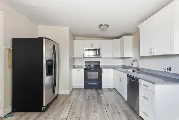 Modern kitchen featuring white cabinetry, stainless steel appliances including a gas range and dishwasher, gray quartz countertops, a stainless steel sink with chrome faucet, and wood-finish flooring throughout the space