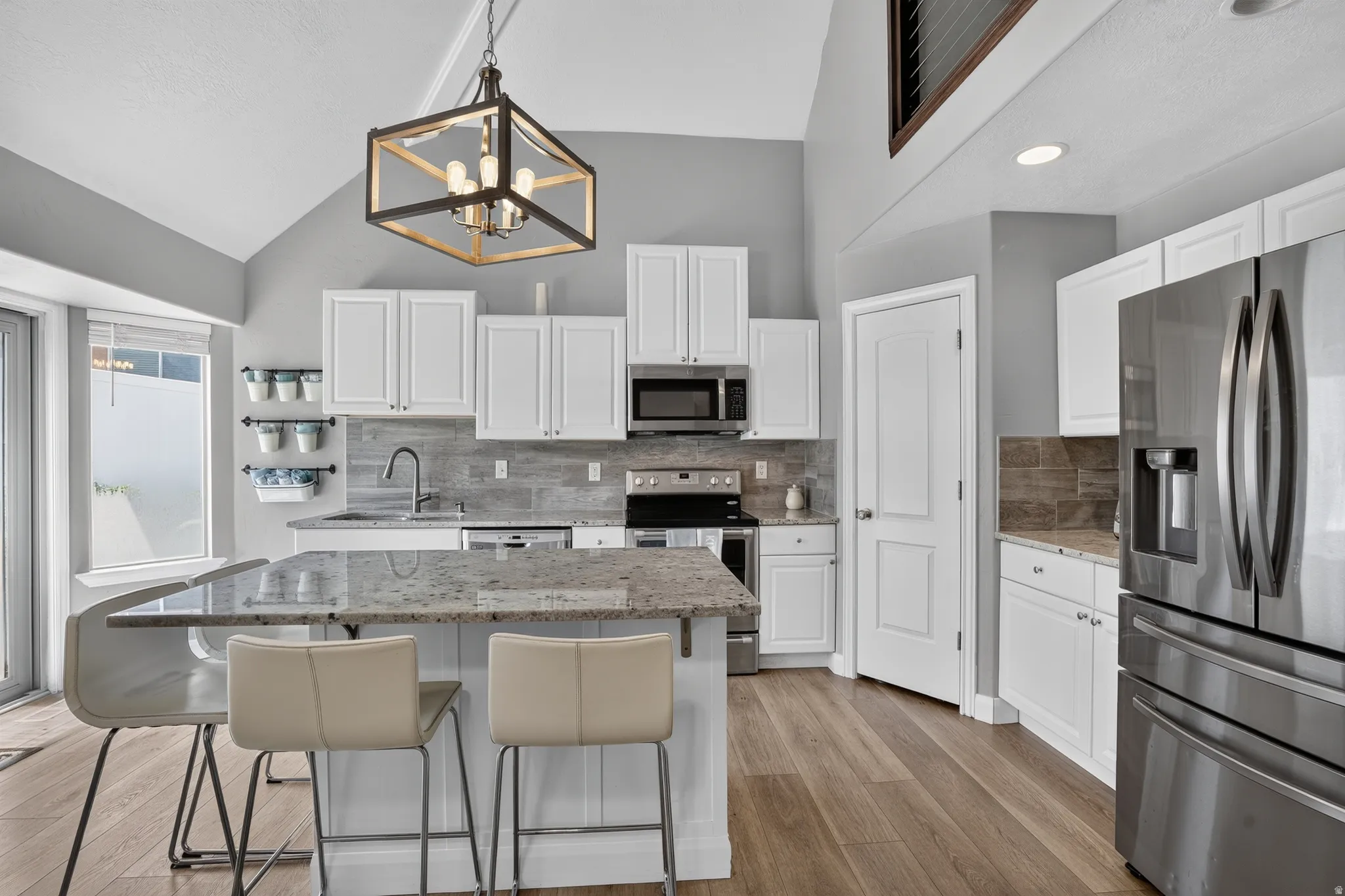 Vaulted ceiling kitchen featuring an expansive island with a light-toned countertop, stainless steel appliances, white cabinetry, wood-finish flooring, and a contemporary chandelier