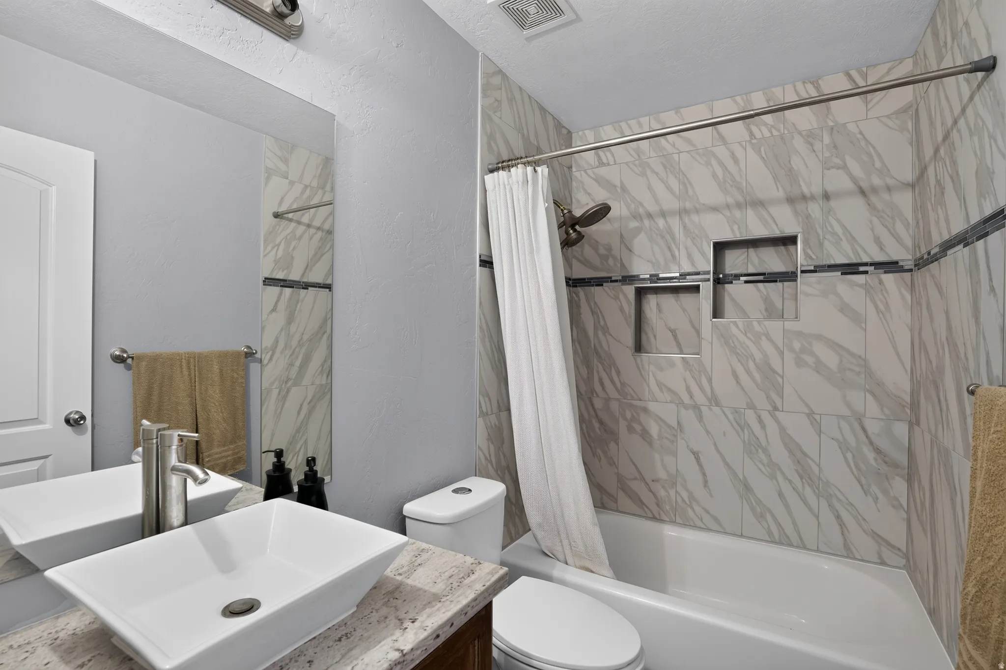 Full bathroom featuring a vessel sink, stone countertop, tub with shower, and tile surround with decorative accent band and integrated niches