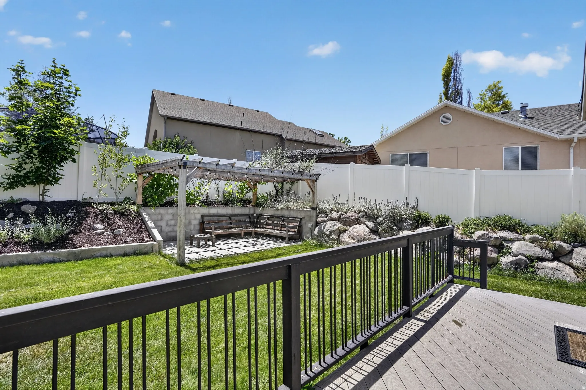 Expansive backyard featuring a composite deck with black metal railing, a manicured lawn, a terraced landscape with natural rock elements, and a pergola-covered patio area with built-in seating