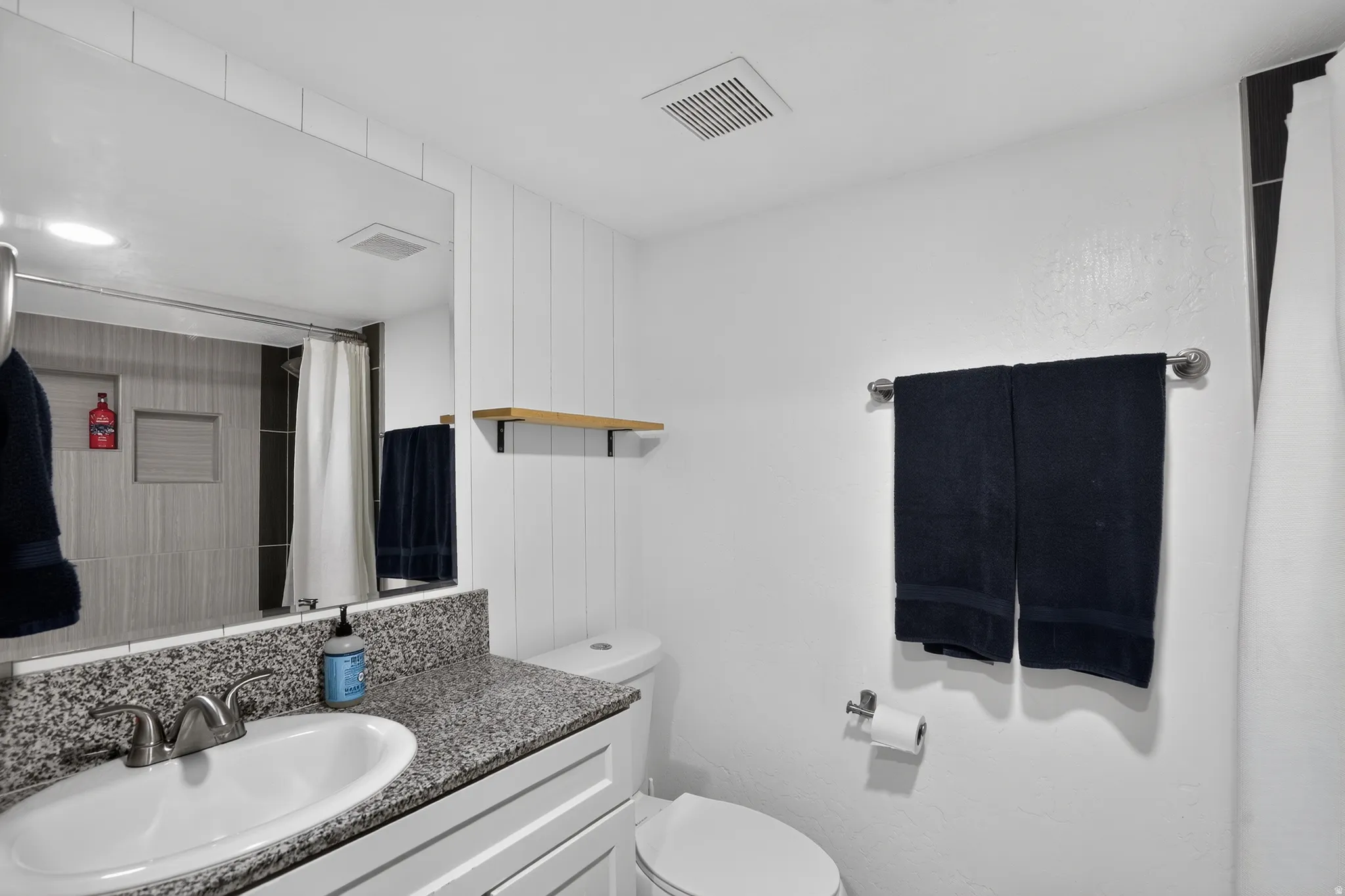 Bathroom featuring a vanity with a speckled countertop and integrated sink, a large mirror, a toilet, and a shower with light-toned tiling