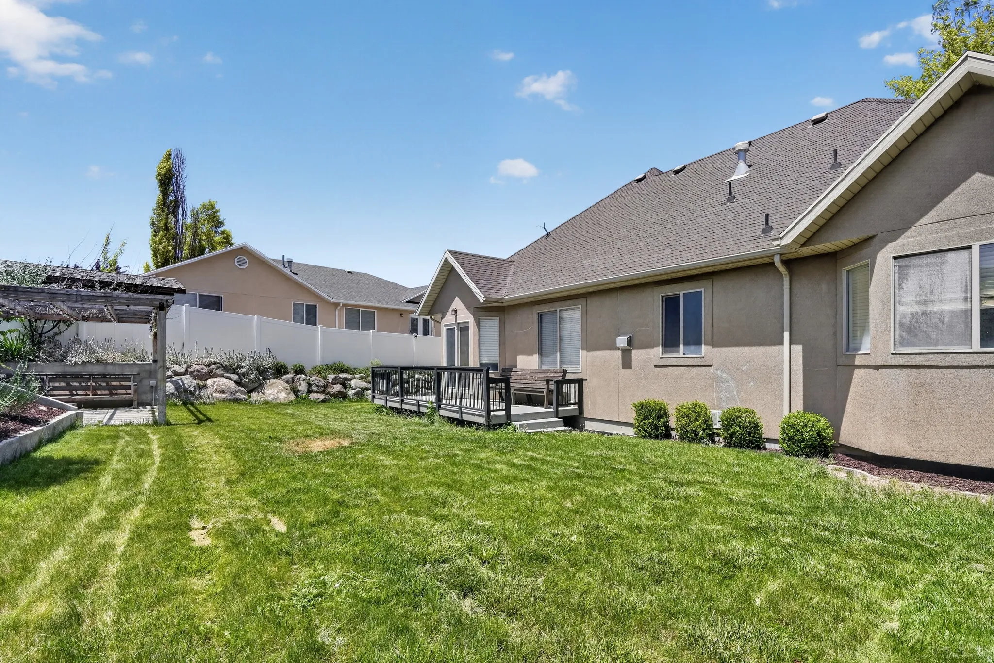 Expansive lawn area featuring a multi-level deck, a wooden pergola structure, a rock retaining wall, mature shrubbery, and a stucco exterior finish