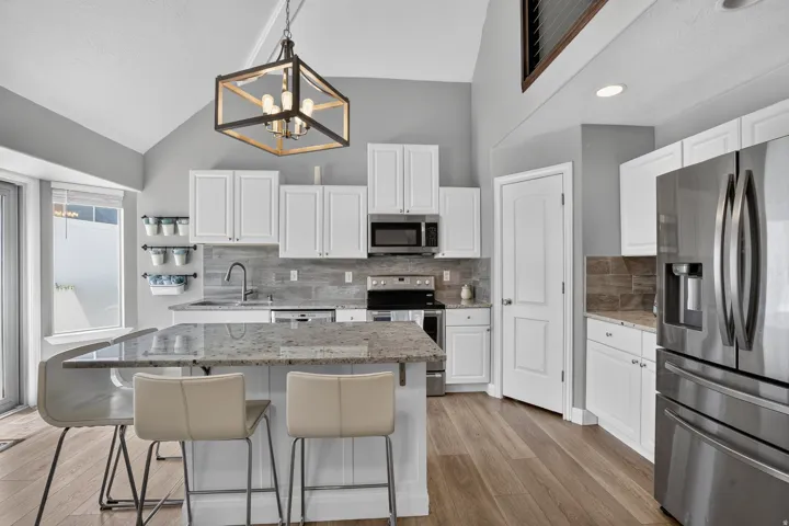 Vaulted ceiling kitchen featuring an expansive island with a light-toned countertop, stainless steel appliances, white cabinetry, wood-finish flooring, and a contemporary chandelier