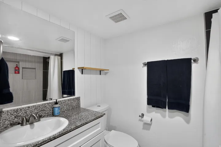 Bathroom featuring a vanity with a speckled countertop and integrated sink, a large mirror, a toilet, and a shower with light-toned tiling
