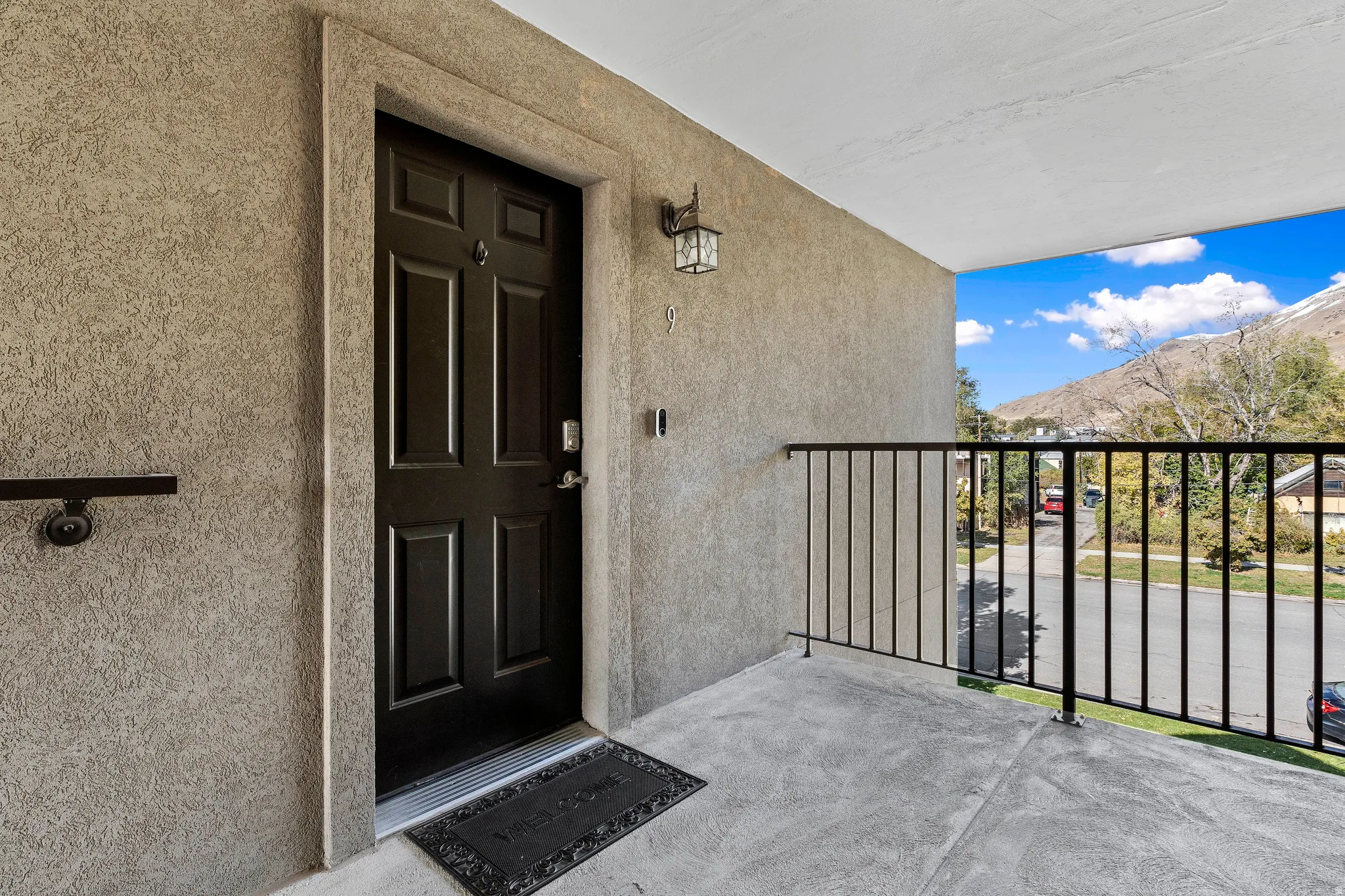 Stucco exterior finish with a paneled entry door, an exterior wall lantern, a metal handrail, and a concrete balcony