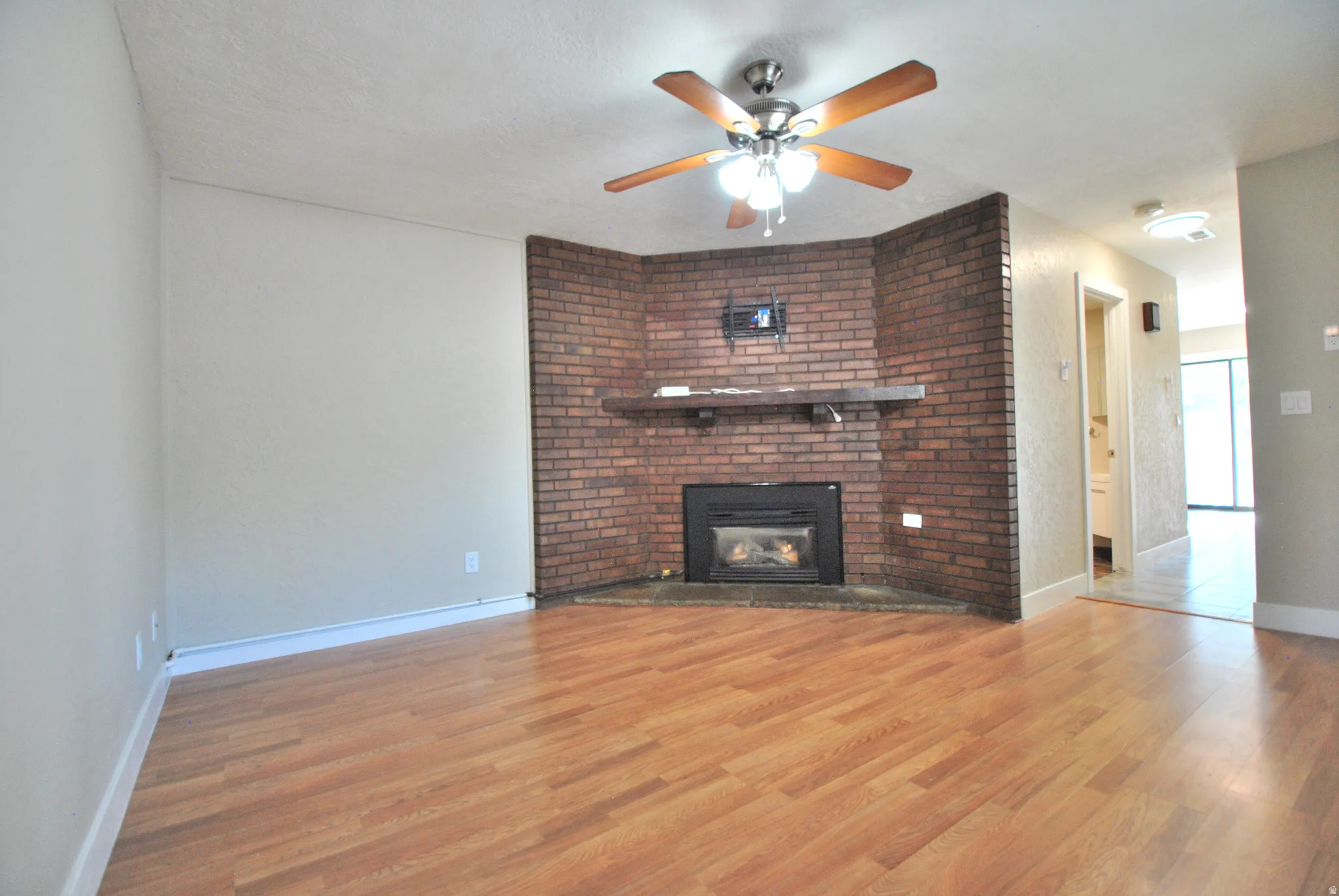 Wood-finish flooring throughout, brick fireplace with a built-in mantel, and a ceiling fan with integrated lighting
