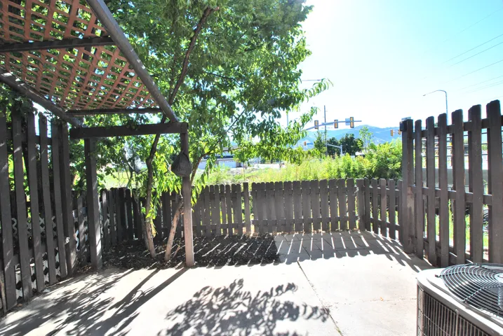 Paved patio area featuring a wooden lattice pergola, surrounded by a wooden picket fence, with mature trees providing natural shade