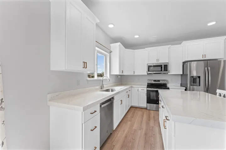 White cabinetry with bronze-finish hardware, stainless steel appliances, light-toned countertops, wood-finish flooring, and recessed lighting