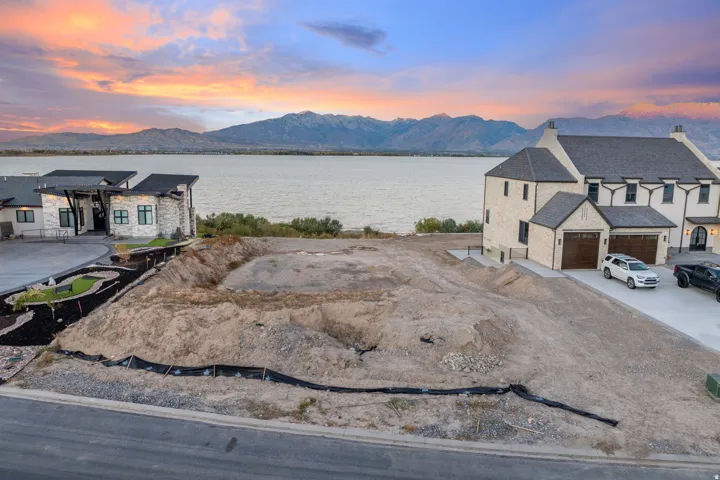Level lot with excavated foundation, bordered by a paved road and established residences, with a prominent body of water and mountain range in the background