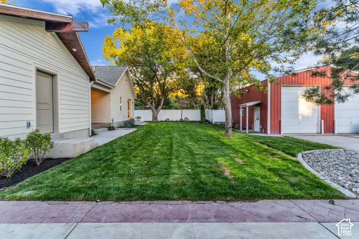 Manicured lawn, mature trees, white privacy fencing, red corrugated metal siding building with two garage doors, and concrete paving