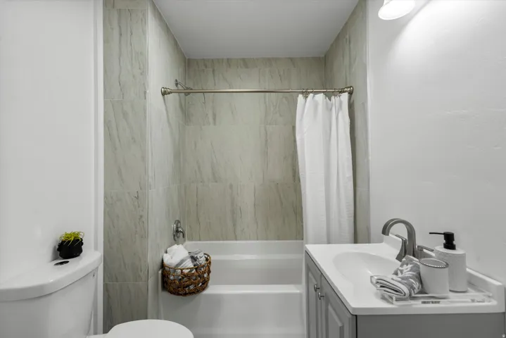 Bathroom featuring a white bathtub, gray tiled shower surround, wall-mounted light fixture, and a vanity with a white countertop and gray cabinetry