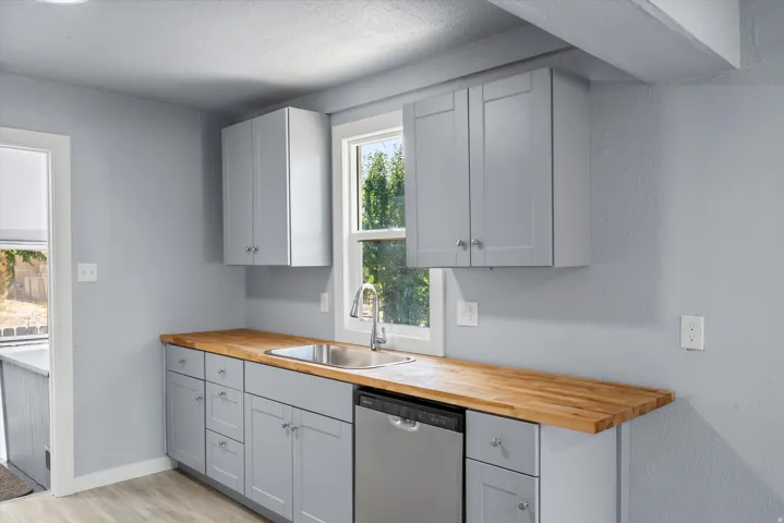Light gray cabinetry with brushed nickel hardware, wood-finish countertops, stainless steel sink, and stainless steel dishwasher