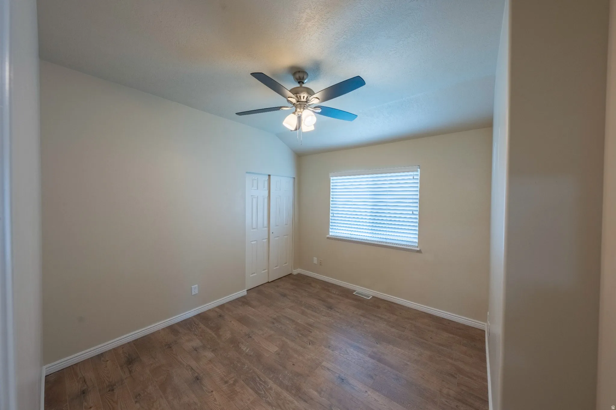 Vaulted ceiling, wood-finish flooring, ceiling fan with integrated lighting, window with blinds, and bi-fold closet doors